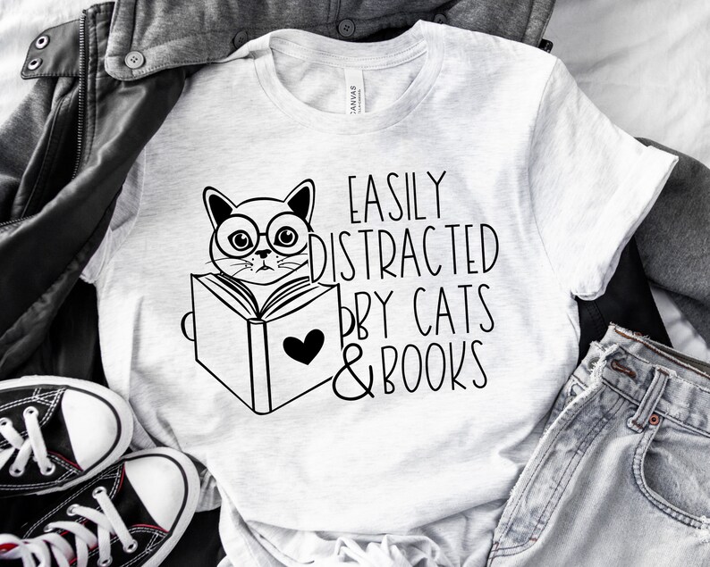 K&ouml;nnte beinhalten: Ein wei&szlig;es T-Shirt mit einer schwarz-wei&szlig;en Illustration einer Katze, die ein Buch liest. Der Text "Easily Distracted by Cats & Books" ist auf das T-Shirt gedruckt.