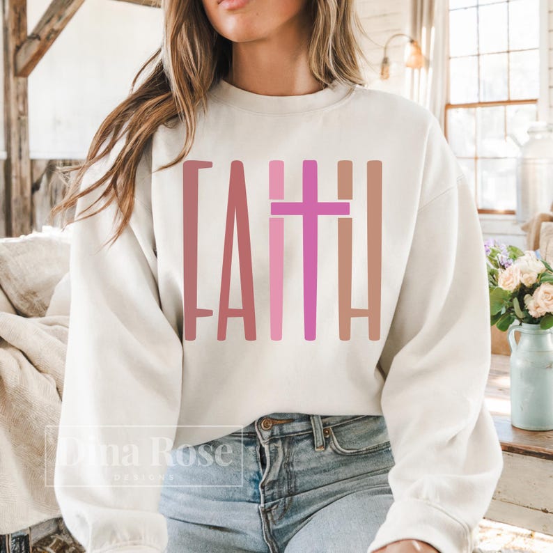 May include: Cream-colored sweatshirt with the word "FAITH" in a vertical, color-blocked design. The letters and cross are in shades of pink and brown. The sweatshirt is a casual, everyday garment.