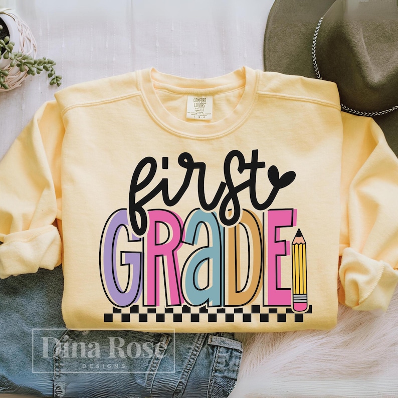 K&ouml;nnte beinhalten: Ein hellgelbes Sweatshirt mit dem Schriftzug "first grade" in einem farbenfrohen Design. Das Wort "grade" ist in Rosa, Blau und Orange gehalten, mit einer gelben Bleistiftgrafik. Unten befindet sich ein schwarz-wei&szlig;es Schachbrettmuster.