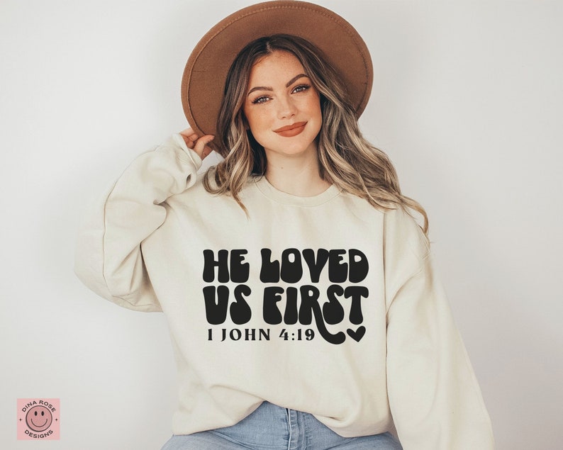 May include: A cream colored sweatshirt with the text "He Loved Us First" in black with a retro font. The text is followed by "1 John 4:19" in a smaller font. A small heart is to the right of the text.