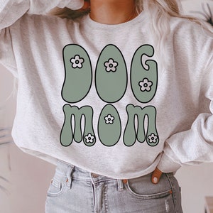May include: A gray sweatshirt with the text "DOG MOM" in green and white. The letters are stylized with a retro 70s font and have small white flowers on them.