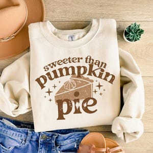 Pode incluir: Uma camisola de cor creme com o texto "Sweeter than pumpkin pie" em letras castanhas. Uma fatia de torta de ab&oacute;bora estilizada em castanho e bege &eacute; apresentada no centro do texto.