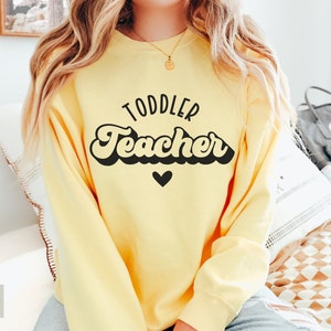 Pu&ograve; includere: Una felpa gialla con il testo "Toddler Teacher" in nero in font retr&ograve; con un piccolo cuore nero sotto il testo.