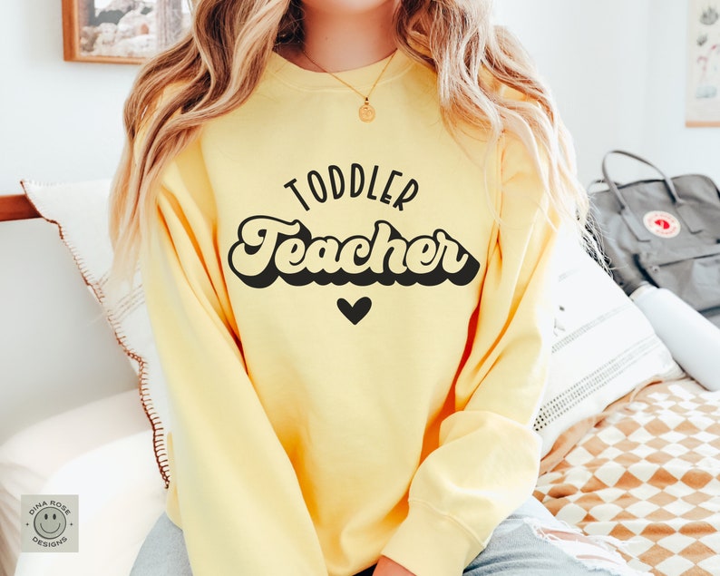 Pu&ograve; includere: Una felpa gialla con il testo "Toddler Teacher" in nero in font retr&ograve; con un piccolo cuore nero sotto il testo.