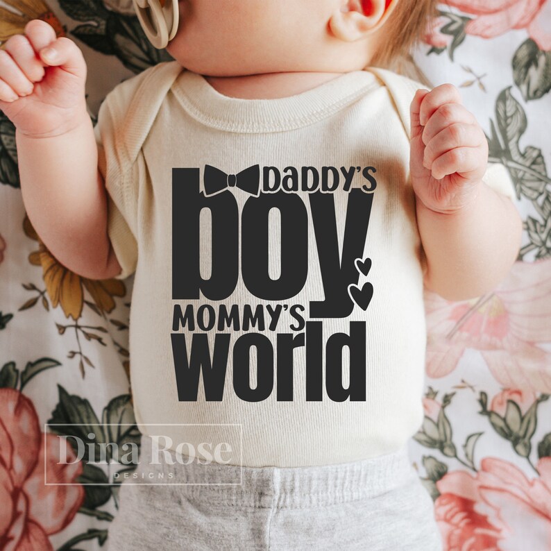 Peut inclure: Body de b&eacute;b&eacute; cr&egrave;me avec l'inscription noire "Daddy's Boy Mommy's World". Le motif comprend un n&oelig;ud papillon et deux c&oelig;urs. Sur un fond floral.