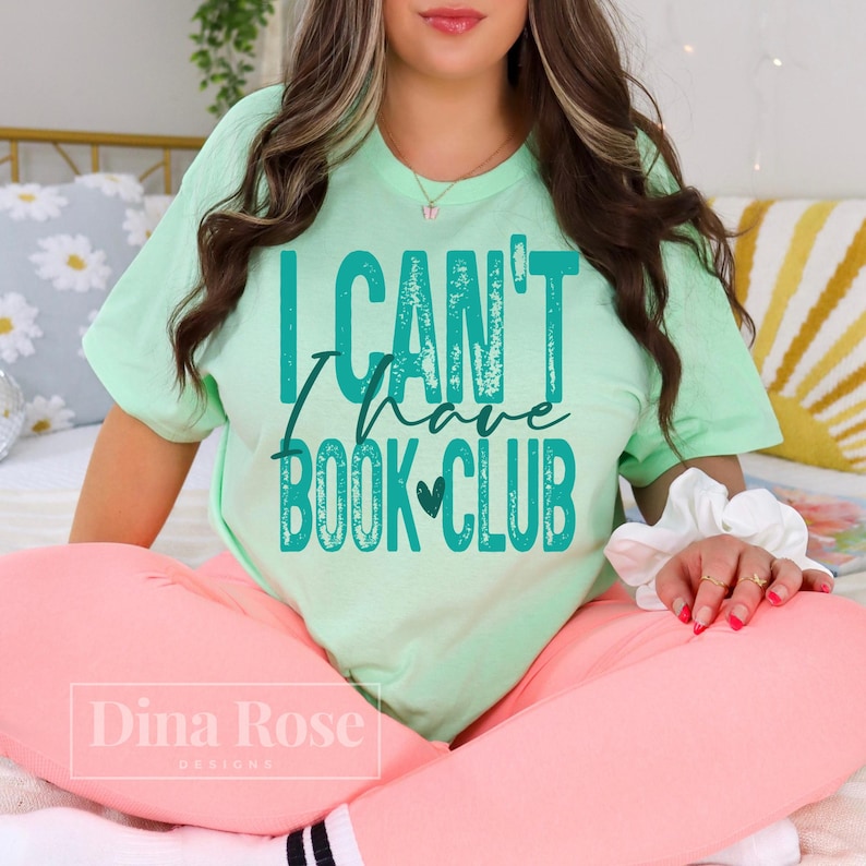 May include: A pale green t-shirt with the teal text "I Can't Have Book Club". The shirt is worn with pink trousers. The image also shows a white scrunchie and a Dina Rose Designs logo.