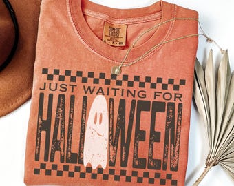 May include: A coral-colored t-shirt with the text "JUST WAITING FOR HALLOWEEN" in dark brown, featuring a ghost graphic. A gold necklace and a brown hat are also visible. The shirt is a Comfort Colors brand.