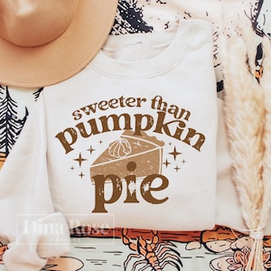 Pode incluir: Uma camisola de cor creme com o texto "sweeter than pumpkin pie" em letras castanhas. O texto &eacute; rodeado por pequenas estrelas e uma ilustra&ccedil;&atilde;o castanha de uma torta de ab&oacute;bora.