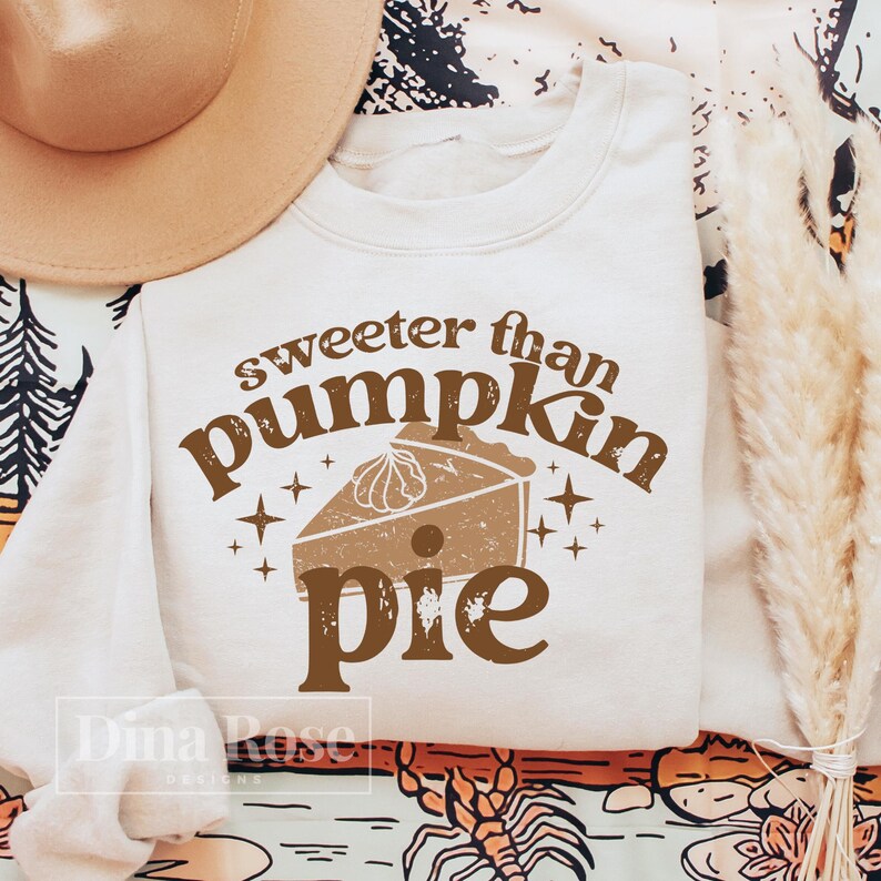 Pode incluir: Uma camisola de cor creme com o texto "sweeter than pumpkin pie" em letras castanhas. O texto &eacute; rodeado por pequenas estrelas e uma ilustra&ccedil;&atilde;o castanha de uma torta de ab&oacute;bora.