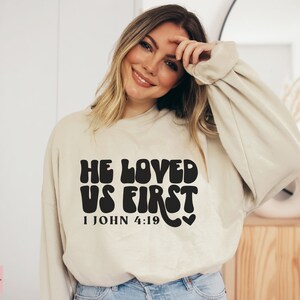 May include: A beige sweatshirt with a black graphic that reads "He Loved Us First 1 John 4:19" in a retro font.
