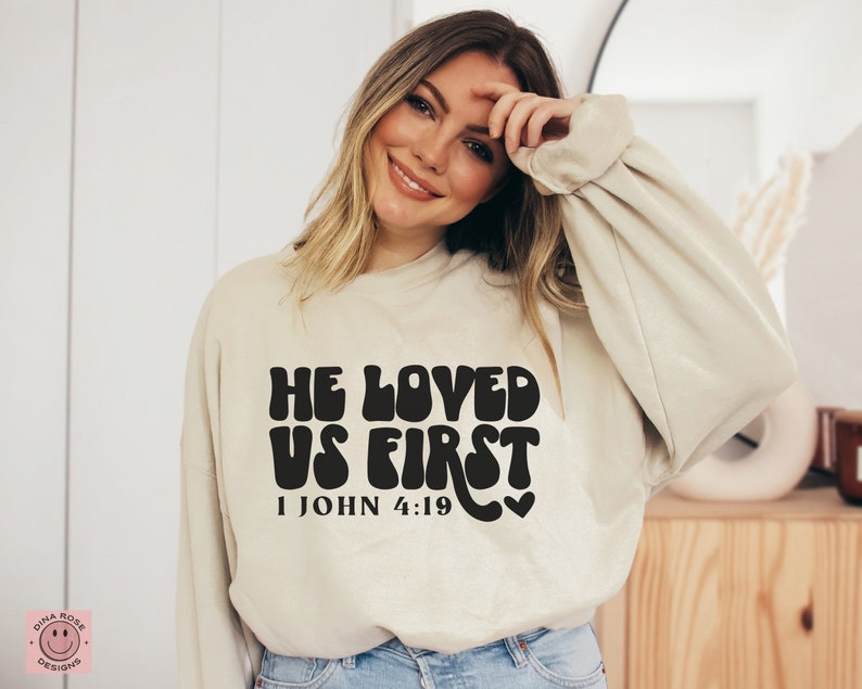 May include: A beige sweatshirt with a black graphic that reads "He Loved Us First 1 John 4:19" in a retro font.