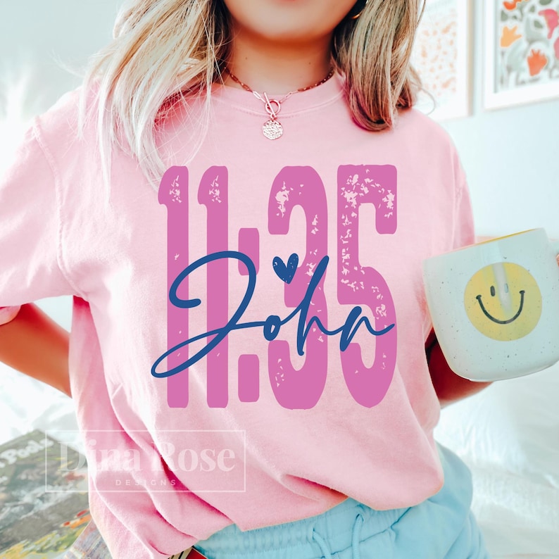 May include: A light pink sweatshirt with the text "11:35 John" in purple and blue. A white mug with a yellow smiley face is held in the hand. The sweatshirt has a crew neck.