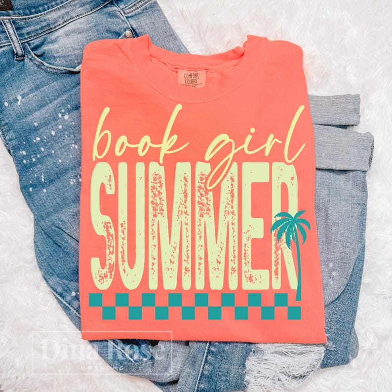 May include: Coral-colored t-shirt with the words "book girl SUMMER" in a light yellow font. A teal palm tree graphic is on the right side. The shirt is folded on top of blue jeans.