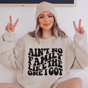 Pu&ograve; includere: Una felpa beige chiaro con una grafica nera che recita "Ain't no family like the one I got" in un font retr&ograve;.