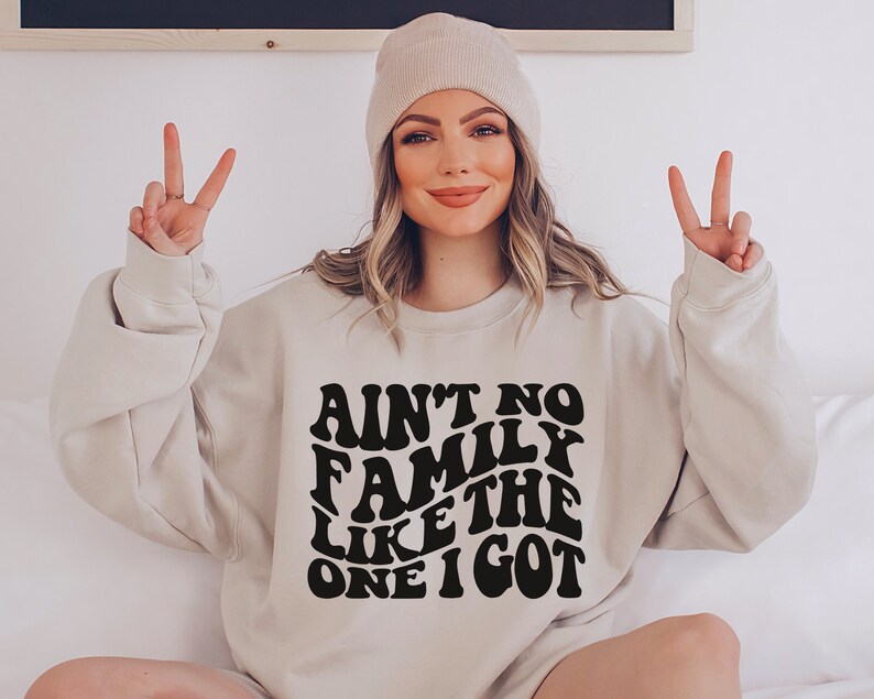 Pu&ograve; includere: Una felpa beige chiaro con una grafica nera che recita "Ain't no family like the one I got" in un font retr&ograve;.