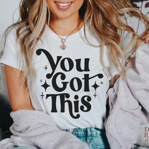 Puede incluir: Camiseta blanca con texto negro que dice "You Got This" en una fuente retro. El texto est&aacute; rodeado de peque&ntilde;as estrellas y signos m&aacute;s.