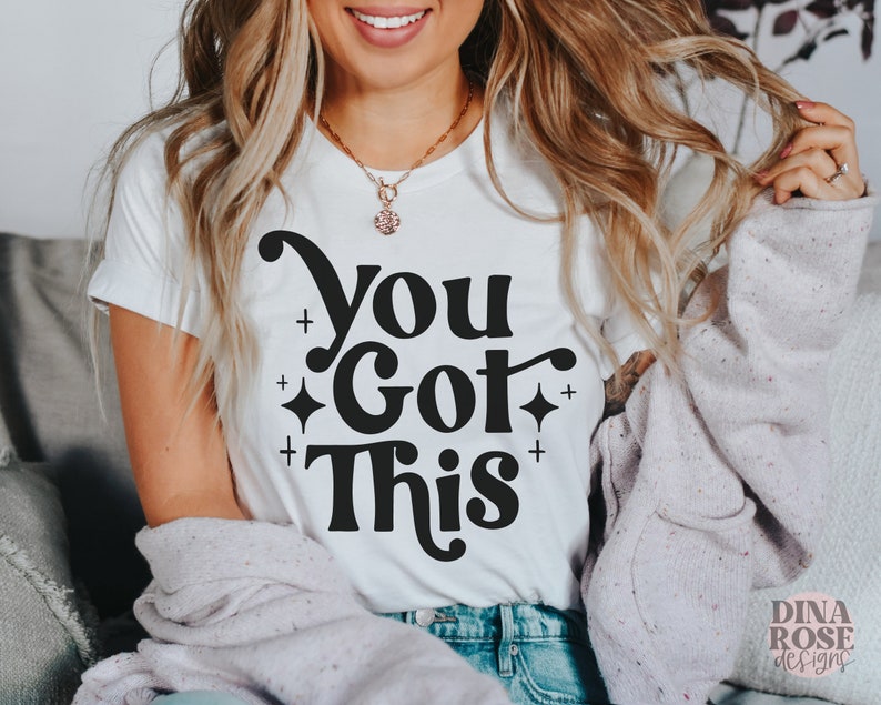 Puede incluir: Camiseta blanca con texto negro que dice "You Got This" en una fuente retro. El texto est&aacute; rodeado de peque&ntilde;as estrellas y signos m&aacute;s.