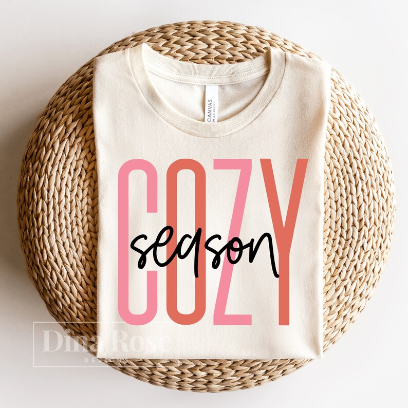 May include: A cream colored t-shirt with the text "COZY season" in pink and orange lettering. The shirt is on a woven straw surface.