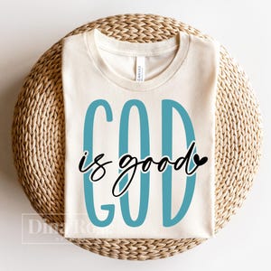 May include: Cream-colored t-shirt with the text "GOD is good" in teal and black script. The shirt is made of a soft material and is displayed on a woven, circular surface. The design is simple and elegant.