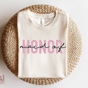 May include: A white t-shirt with the text "maid of honor" in pink letters. The shirt is folded and displayed on a woven surface.