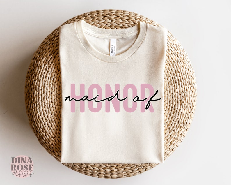 May include: A white t-shirt with the text "maid of honor" in pink letters. The shirt is folded and displayed on a woven surface.