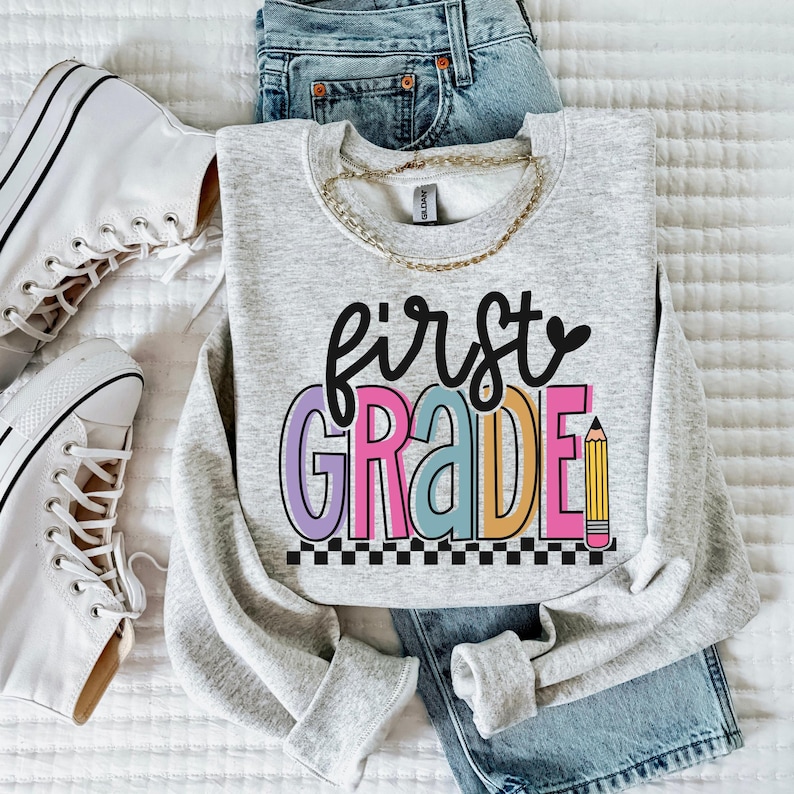 K&ouml;nnte beinhalten: Ein hellgraues Sweatshirt mit den Worten "first grade" in bunten Buchstaben und einer Bleistiftgrafik. Das Sweatshirt wird mit einer blauen Jeans, einer goldenen Halskette und wei&szlig;en Turnschuhen kombiniert.