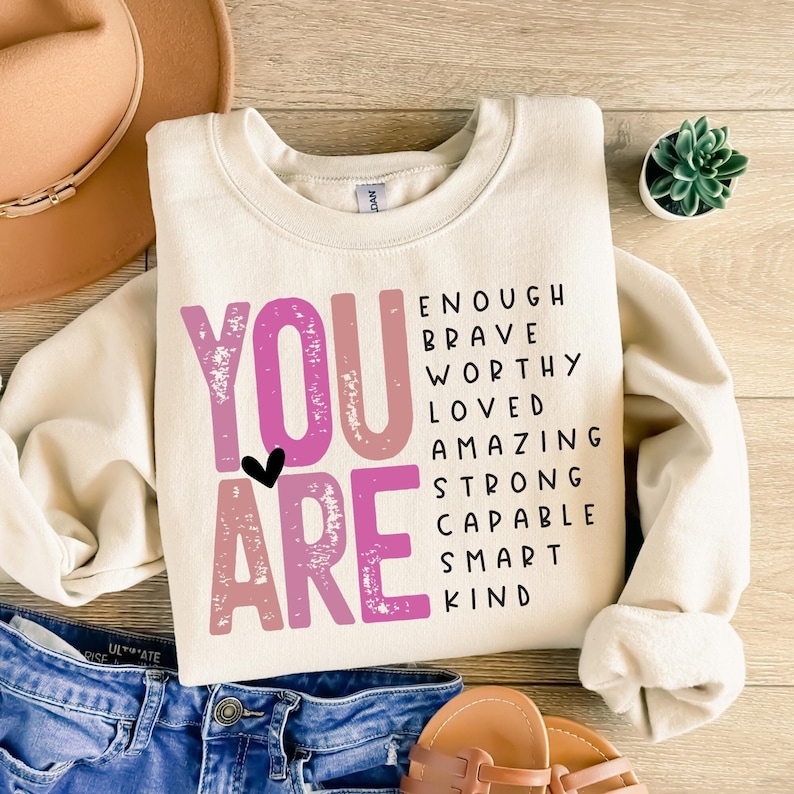 May include: A cream-colored sweatshirt with the words "YOU ARE" in pink and white letters. The words are surrounded by a list of positive affirmations, including "enough", "brave", "worthy", "loved", "amazing", "strong", "capable", "smart", and "kind".