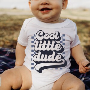 May include: A white baby onesie with a blue and white checkered background and the text "Cool little dude" in a retro font.