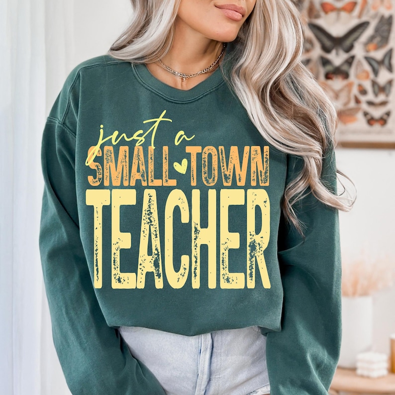 May include: A dark green sweatshirt with the text "just a SMALL TOWN TEACHER" in yellow and orange lettering. The sweatshirt is a crew neck style and appears to be made of a soft material. The overall design is simple and casual.