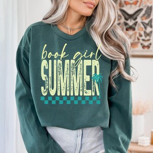 May include: Dark teal sweatshirt with the words "book girl SUMMER" in a yellow, distressed font. A palm tree graphic is on the right side of the word "SUMMER". A teal and white checkered pattern is at the bottom of the design.