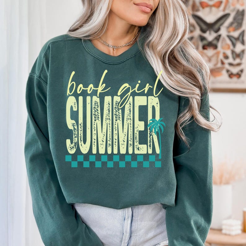 May include: Dark teal sweatshirt with the words "book girl SUMMER" in a yellow, distressed font. A palm tree graphic is on the right side of the word "SUMMER". A teal and white checkered pattern is at the bottom of the design.