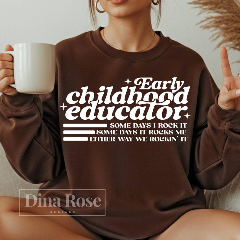 以下が含まれることがあります： 「Early Childhood Educator」の白文字がプリントされた茶色のスウェットシャツ。「Some days I rock it」、「Some days it rocks me」、「Either way we rockin' it」のフレーズも含まれています。白いマグカップが左手に持たれています。