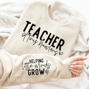 May include: A beige sweatshirt with the text "TEACHER of tiny humans" on the front and "HELPING little minds GROW" on the sleeve.