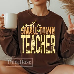 May include: A brown sweatshirt with the text "just a SMALL TOWN TEACHER" in yellow and orange. The sweatshirt has a crew neck and long sleeves. A white mug is held in the left hand.
