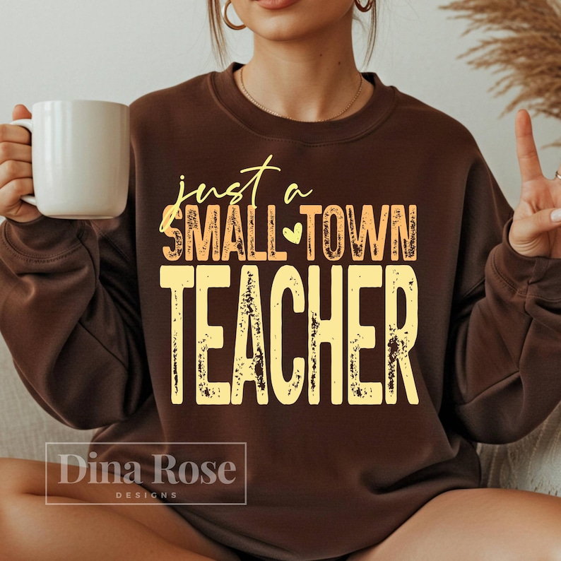 May include: A brown sweatshirt with the text "just a SMALL TOWN TEACHER" in yellow and orange. The sweatshirt has a crew neck and long sleeves. A white mug is held in the left hand.