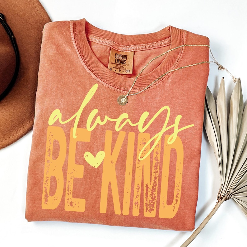 May include: Orange Comfort Colors t-shirt with the words "always be kind" in yellow and orange. A gold necklace with a small pendant rests on the shirt. A brown hat and dried plant are in the background.