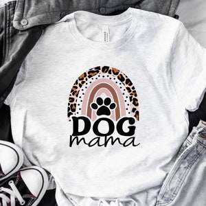 May include: White t-shirt with a rainbow design featuring a paw print in the center. The rainbow is made of pink, orange, yellow, green, blue, and purple stripes. The text "DOG mama" is written below the rainbow.