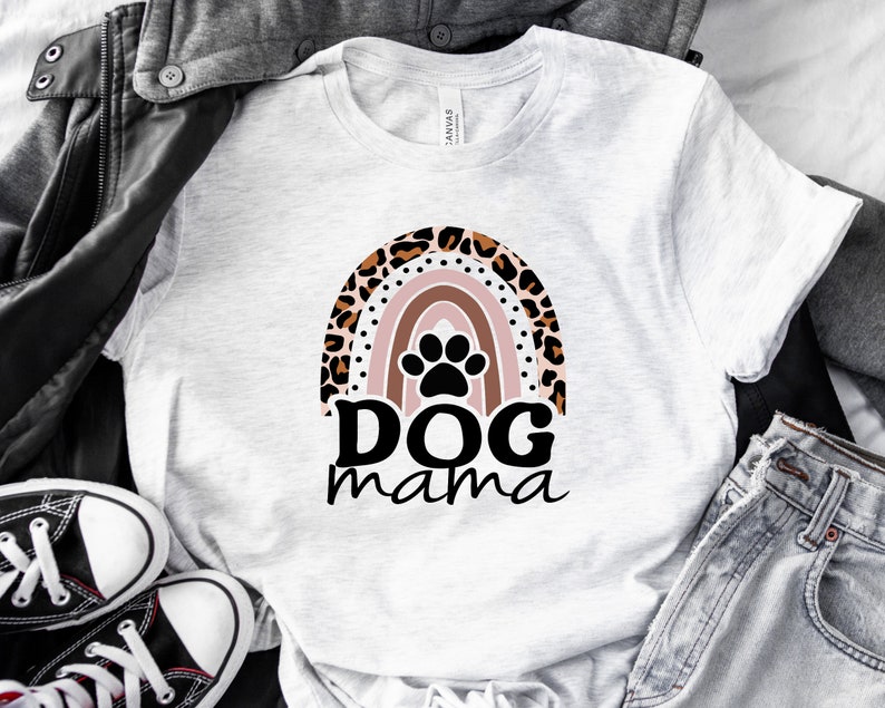 May include: White t-shirt with a rainbow design featuring a paw print in the center. The rainbow is made of pink, orange, yellow, green, blue, and purple stripes. The text "DOG mama" is written below the rainbow.