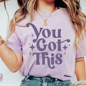 Puede incluir: Una camiseta morada con el texto "You Got This" en una fuente retro. El texto est&aacute; en color morado oscuro y est&aacute; rodeado de peque&ntilde;as estrellas blancas.
