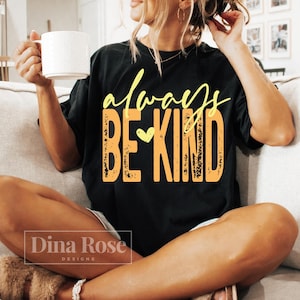 K&ouml;nnte beinhalten: Schwarzes T-Shirt mit den Worten "always BE KIND" in gelber und orangefarbener Schrift. Das Shirt hat eine Herzgrafik. Das Dina Rose Designs-Logo befindet sich unten. Die Person h&auml;lt einen wei&szlig;en Becher.