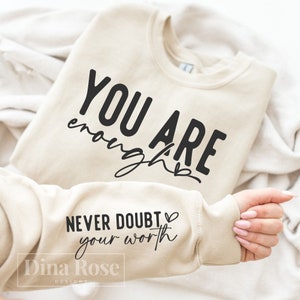 May include: A beige sweatshirt with the text "YOU ARE enough" on the front and "NEVER DOUBT your worth" on the sleeve. The text is in black.