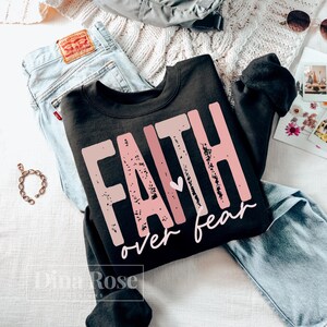May include: A black sweatshirt with the words "FAITH over fear" in distressed pink lettering. The sweatshirt is paired with light-wash denim jeans, a gold bracelet, and sunglasses, creating a casual, everyday aesthetic.