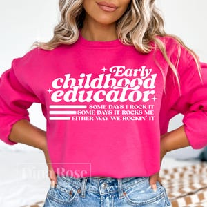 以下が含まれることがあります： 鮮やかなピンク色のスウェットシャツ。白文字で「Early Childhood Educator」と書かれています。その下に「SOME DAYS I ROCK IT, SOME DAYS IT ROCKS ME, EITHER WAY WE ROCKIN' IT」と書かれています。クルーネックのデザインです。