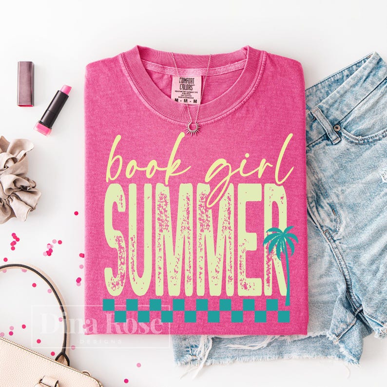 May include: A hot pink t-shirt with the words "book girl SUMMER" in a distressed, light green font. A palm tree graphic is on the right side of the word "SUMMER". The shirt is folded and next to denim shorts.