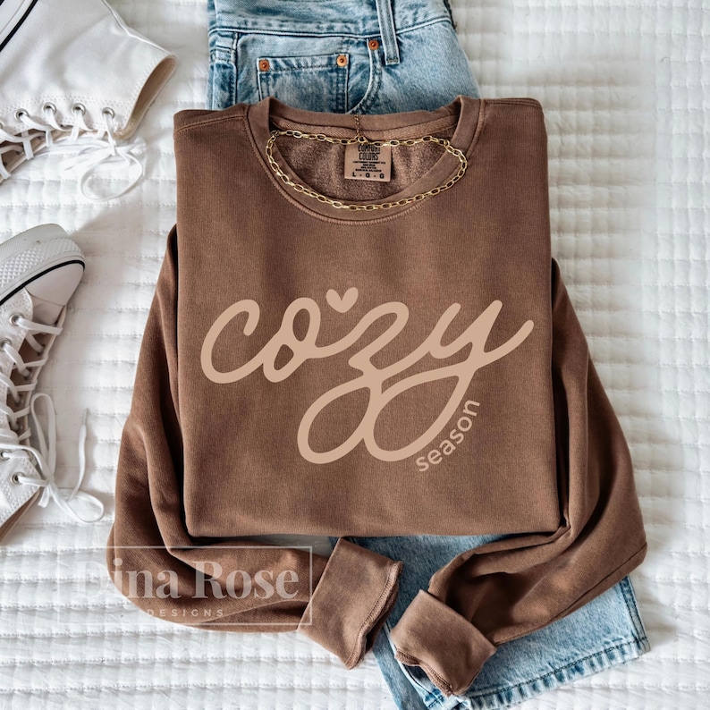 May include: A brown sweatshirt with the words "cozy season" in a cursive font. The sweatshirt is paired with blue jeans, a gold chain necklace, and white sneakers. The image is taken from a top-down perspective.
