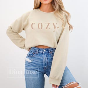 May include: A beige sweatshirt with the word "COZY" printed across the chest in a light brown color. The word "COZY" has a small heart at the end. The model is wearing blue jeans.
