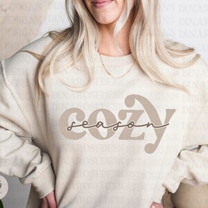 May include: A beige sweatshirt with the text "cozy season" in brown lettering. The text is in a stylized font with a curved "C" and "Y".
