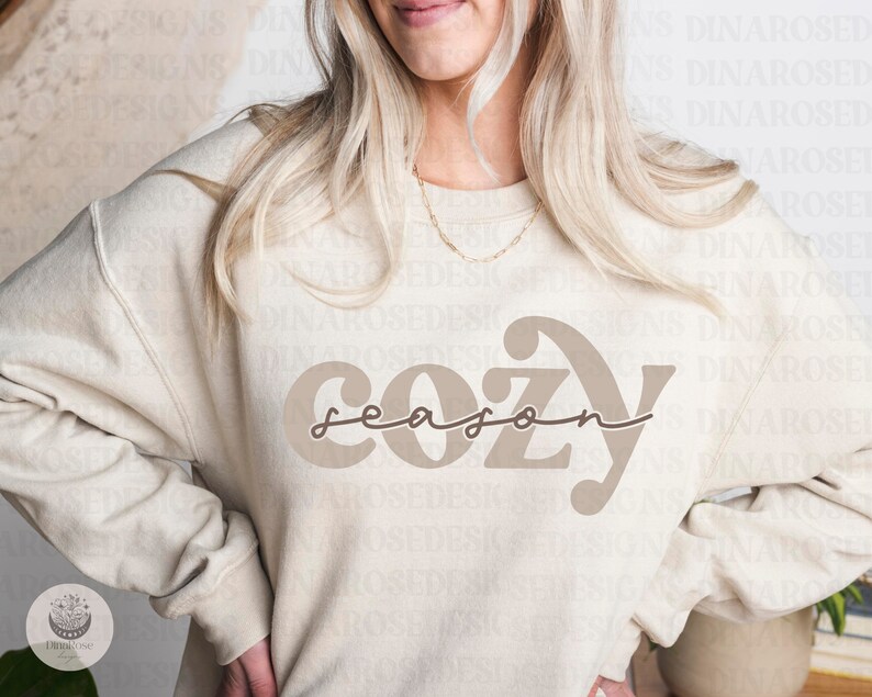May include: A beige sweatshirt with the text "cozy season" in brown lettering. The text is in a stylized font with a curved "C" and "Y".