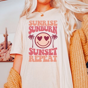 May include: A white t-shirt with a pink and peach graphic design that says "Sunrise Sunburn Sunset Repeat" with a smiley face with hearts for eyes and palm trees on either side.