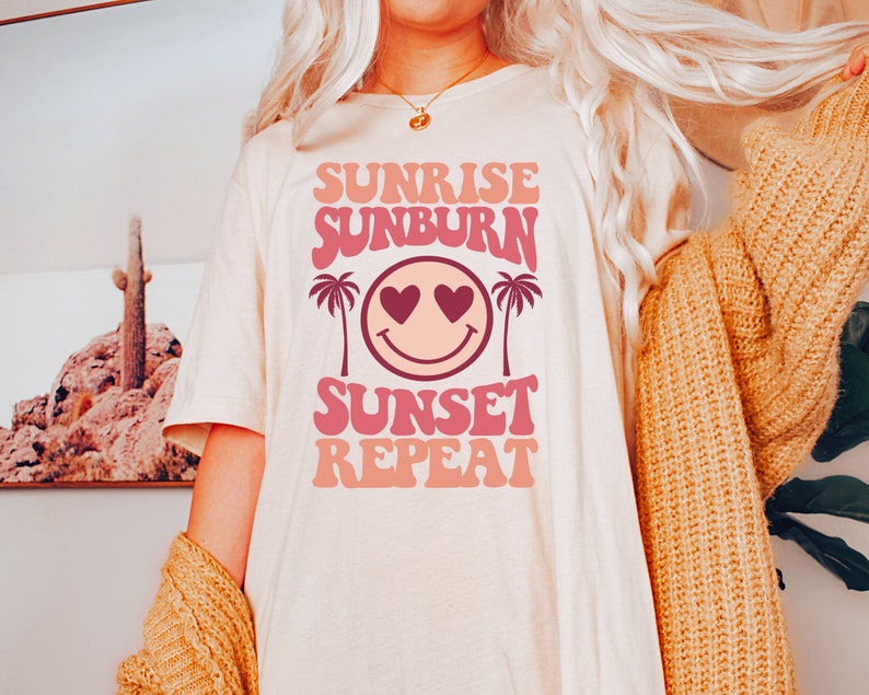 May include: A white t-shirt with a pink and peach graphic design that says "Sunrise Sunburn Sunset Repeat" with a smiley face with hearts for eyes and palm trees on either side.